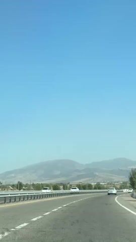 The video shows a view from a moving vehicle on a highway. The sky is clear and blue. In the distance, there are mountains and trees. There are several cars on the highway, including a white truck, a white sedan, and a white SUV. The highway has a guardrail and white lines marking the lanes. The video is in landscape orientation.