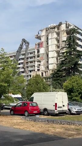 A demolition crane is actively dismantling a multi-story building. The crane, with its long arm extended, is shown impacting the upper floors of the structure, causing debris to fall. The building itself appears to be a concrete and possibly brick structure, with visible floors and window openings. In the foreground, several vehicles are parked, including a red car, a white van, and a silver car, suggesting the demolition is taking place in an urban or suburban area. Trees are visible behind the vehicles and around the building, indicating an outdoor setting. The sky is clear and blue. The action is focused on the controlled destruction of the building.