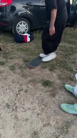 The video shows a person in black pants and white sneakers attempting to do a trick on a skateboard. The skateboard is on the ground, and the person is standing with one foot on it. The person jumps, and the skateboard flips up. Another person wearing green shoes is standing to the right. A black car is parked in the background, with its door open, and a bag is on the ground next to it. The ground is covered with dirt and grass.