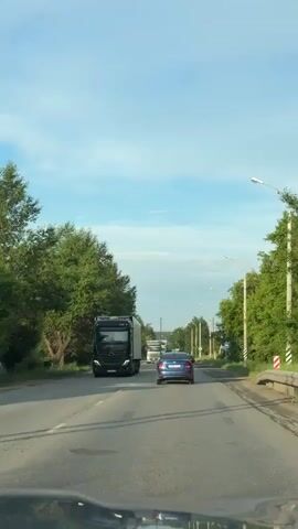 A blue sedan is driving on a two-lane road. A black truck is on the left side of the road. There are trees on both sides of the road. The sky is blue with some clouds. The camera is inside a car looking out the windshield.