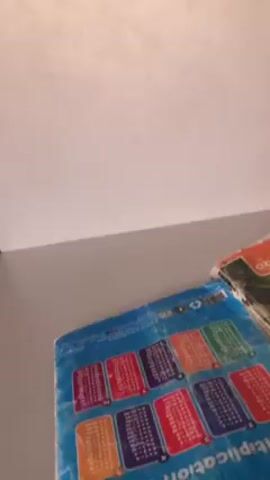 The video shows the inside of a library. There are study carrels with white tops and white privacy partitions. A green plastic chair is in front of one of the carrels.  A couple of books are on the desk of one carrel.  A bookshelf filled with books is in the background. In one shot, a person is sitting at one of the carrels with their legs extended onto the desk of another carrel and reading something. The lighting in the room appears to be bright.