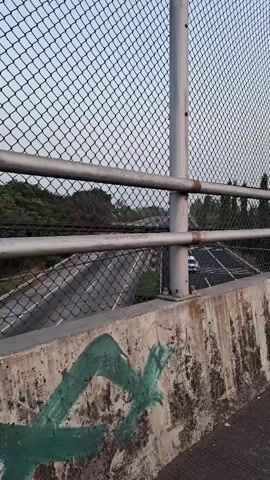 The video begins with a view of a concrete bridge railing with graffiti, then pans right to show a chain-link fence and a road below. The perspective shifts to show a street scene with several motorcycles riding along a road, some with multiple passengers. The sky is overcast and appears to be either early morning or late evening. Trees and other vegetation are visible in the background. The video ends with a close-up shot of two people on a motorcycle.