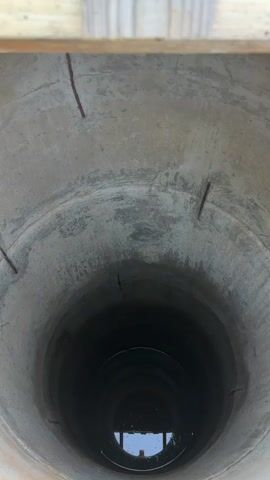 This is a vertical, downward-facing shot inside a concrete well. The well walls are rough and show signs of age and wear, with some cracks and rust stains visible. Towards the top, a wooden beam is partially visible. The well narrows as it goes down, becoming darker and eventually showing a pool of dark water at the bottom. The water reflects some light, suggesting a light source from above or the side. Small bubbles and ripples are visible on the water's surface. The overall impression is of a deep, potentially abandoned, structure.