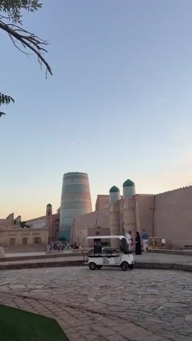 A white golf cart is parked in an open paved area. In the background, there are several old buildings with distinctive architecture, including a tall tower with a patterned exterior and two domed structures. The sky is a clear, pale blue. A few people are visible walking around the buildings. A tree branch extends into the frame from the upper left corner.