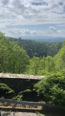 The video shows a view of a lush, green valley with mountains in the distance under a partially cloudy sky. The view is framed by trees and bushes in the foreground, with a stone wall and a section of what appears to be steps or pavement. The camera slowly pans up to show more of the sky and mountain range.