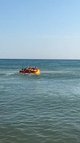 A yellow and red inflatable tube boat with at least four people is being pulled across the blue ocean by a jet ski. The sky is clear and blue. The water is calm with small waves.