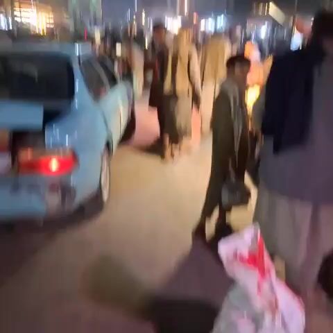 The video depicts a nighttime street scene filled with people and vehicles. White banners with Arabic script are strung across the street, and colorful lights illuminate a building in the background. Several cars, including a light blue sedan, are parked or moving through the crowd. People are seen walking, talking, and standing around, some wearing traditional clothing. The overall atmosphere suggests a public gathering or event.