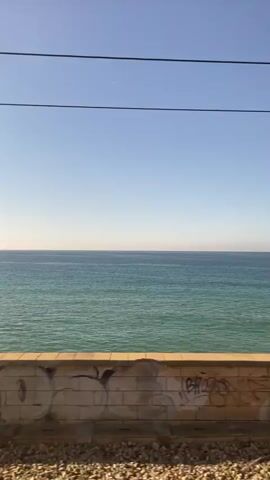 A video captures a view from a moving train window overlooking the ocean. The camera focuses on the sea, the sky, and elements of the train's surroundings. In the foreground, a low wall is visible, adorned with graffiti. Beyond the wall, the turquoise sea stretches out to the horizon under a clear blue sky. Power lines stretch across the top of the frame. The video shows a few man-made structures near the shore, including a wooden walkway with railings and a signpost. The train appears to be traveling along a coastal route, offering glimpses of the seaside environment. There are no people visible in the video.