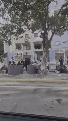 The video shows the exterior of a damaged apartment building. The facade of the building is heavily damaged, with sections of the walls and balconies collapsed. Debris litters the ground in front of the building. Several people are gathered outside, some sitting on furniture that appears to have been removed from the apartments. A refrigerator and mattresses are also visible on the sidewalk. The overall scene suggests a recent disaster, likely an earthquake or explosion, that has caused significant structural damage to the building and displaced its residents. The environment appears to be a residential urban area, with a street in the foreground.