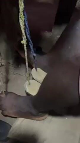 A video shows the legs and feet of a dark-skinned person. They are operating a wooden treadle connected by ropes to an unseen mechanism. The person is barefoot, and their feet move rhythmically on the treadle. The ground appears to be dirt, and there are other objects, including what looks like a shoe, partially visible. The lighting is dim, and the focus is on the person's feet and the treadle.