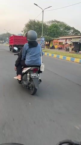 The video shows a view from behind a motorcycle traveling on a paved road. On the motorcycle are two people. The person in front is wearing a blue jacket and a helmet. The person in the back is wearing black pants and a light-colored shoe. They are both wearing helmets. To the left of the motorcycle, a large red dump truck is driving in the same direction. The environment appears to be urban or suburban, with buildings and trees visible on the right side of the road. The sky is overcast or hazy. The motorcycle has a license plate.