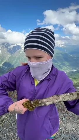 The video shows a person wearing a purple jacket, a striped beanie, a face mask, and colorful shorts. The person is standing on a rocky mountain peak, holding a stick above their head. The background features snow-capped mountains and a valley. The person then lowers the stick and begins to walk down the mountain, turning the camera to show the expansive mountain landscape. The weather is clear with some clouds.