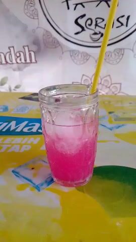 A close-up shot of a clear glass filled with a pink beverage and ice cubes. A yellow straw extends from the top of the glass. The glass sits on a table with a yellow surface that has some water droplets and a green element on the right side. In the background, there is a white sheet with some text and designs.