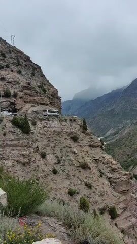 A white vehicle is driving on a road carved into the side of a rocky, mountainous terrain. The road is narrow and winding, with steep drop-offs on one side. The mountains in the background are shrouded in mist and clouds. The vegetation on the slopes is sparse, consisting of scrubby bushes and a few small trees. The sky is overcast, suggesting a cloudy or rainy day.
