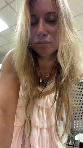 A woman with long blonde hair and a nose ring is leaning forward, her hands near her face. She is wearing a light pink, ruffled dress and several necklaces. Her right hand is raised towards her face, and her fingers are slightly blurred, indicating movement. She appears to be in an indoor setting with a tiled ceiling and a metal chair visible to her right. The lighting is overhead and somewhat dim.