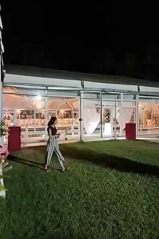 The video captures an outdoor evening scene at a venue with a large, white tent structure. The tent has glass walls, revealing tables and decorations inside, suggesting an event is taking place. Outside the tent, a grassy lawn is visible, with some trees and foliage.

In the foreground, a person is walking across the grass. This individual is wearing a dark top and striped pants. Further in the background, a man in a white shirt and shorts is walking towards the tent. Another person, wearing a dark jacket and jeans, is walking in the opposite direction on the lawn. Inside and around the tent, other individuals are present, some near tables and others walking. The area is illuminated by artificial lights, casting shadows on the grass. Two large red cylindrical objects are placed on the lawn near the tent. The overall atmosphere appears to be that of a social gathering or celebration.