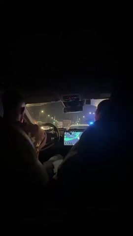 This is a first-person view from the backseat of a car at night. The driver is visible in the front left, with their hands on the steering wheel. A person is sitting in the front right passenger seat, their face turned away from the camera. The car's dashboard and infotainment screen are illuminated. Outside the car, streetlights and the headlights of other vehicles are visible through the windshield, indicating a city environment. The video also shows a brief glimpse through the side window, revealing more traffic and streetlights. The overall impression is that of being in a car moving through a city at night.