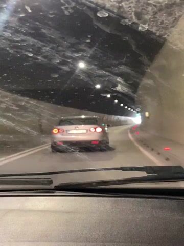 The view from inside a car driving through a tunnel. The tunnel is lit by overhead lights and has a car ahead. The walls are rough and the ceiling is dark. The windshield wipers are visible at the bottom of the frame.
