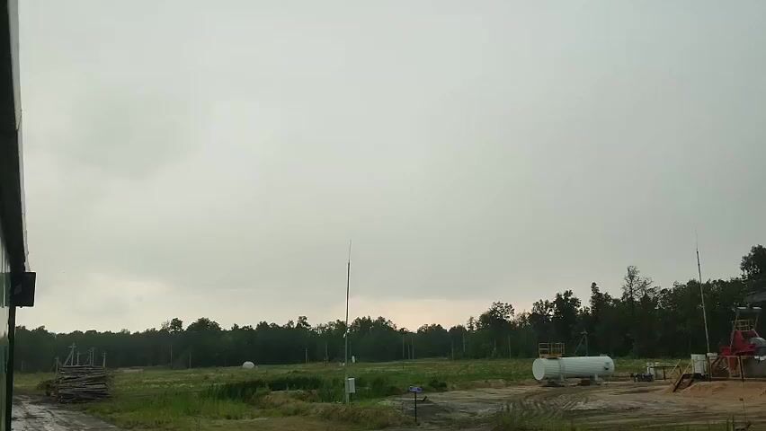 This media file captures an outdoor scene under a cloudy sky, likely during the daytime due to the overall brightness. The setting appears to be a somewhat industrial or rural area, with a line of trees forming the background. In the foreground and mid-ground, several objects are visible. To the left, a dark structure, possibly part of a building or vehicle, encroaches on the frame. Further into the field, there are stacks of lumber, a large white cylindrical tank, and some industrial equipment, including what looks like part of a structure with red elements. There are also tall, thin poles with what might be antennae or lighting fixtures scattered across the landscape, and a white spherical object, perhaps a covered tank or storage. The ground is uneven and appears to be dirt or gravel, with patches of grass. The sky is overcast, suggesting a cloudy day, potentially with overcast weather. The overall impression is one of a functional, perhaps active, outdoor site with hints of natural surroundings. There is no visible text in the media file.