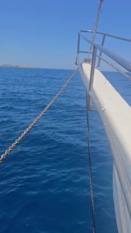 The video begins from the perspective of someone on a boat, looking forward. A thick metal chain extends from the bow of the boat into the deep blue ocean. The water is calm with gentle waves. In the distance, a coastline with mountains is visible under a clear blue sky. The camera pans slightly to the left, revealing more of the ocean and the distant land.