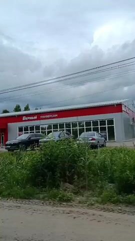 The video shows a side view of a red and white building with large windows and signs. The signs are in Russian and appear to indicate a supermarket or department store. Several cars are parked in front of the building, and people can be seen walking in and out. The building is surrounded by greenery, including trees and grass. The sky is cloudy. The camera is moving, suggesting that the video was taken from a vehicle.