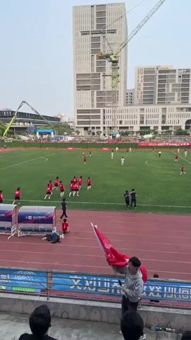 A wide shot from stadium seats shows a soccer game in progress, which ends with one team in red shirts celebrating a victory. The winning team runs onto the field, some taking off their shirts as they approach the apparent top scorer with his arms spread wide, and the teammates proceed to dogpile him. On the sidelines, spectators watch the game and celebrate with a flag. In the background are tall, nondescript buildings and construction equipment. The weather appears to be overcast.