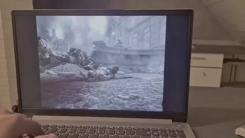 The video shows a laptop screen displaying a video game. The game appears to be a war scene with soldiers and buildings. In the foreground of the game, there is a fallen soldier lying on the ground. In the background, there are more soldiers and a building with smoke coming out of it. A hand is visible on the keyboard of the laptop, suggesting someone is playing the game. The overall scene is monochrome, giving it a gritty, realistic feel.
