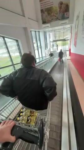 From a first-person perspective, a shopping cart full of glass bottles of cooking oil is moving on a moving walkway in a large, well-lit supermarket. Another man wearing a black jacket is standing in his own shopping cart ahead of the camera, also on the moving walkway. In the distance, two people are walking away through the exit doors of the supermarket. Large windows on the left side of the walkway allow natural light to illuminate the space. A large advertisement is visible on the wall above the walkway. Red accents are visible along the wall to the right.