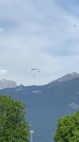 The video shows a paraglider floating in the sky, with a mountainous background. The sky is a pale blue with some clouds. The mountains are green with patches of snow near their peaks. Two trees are visible in the foreground, and a light pole can be seen between them. The paraglider is blue and red, and a person is suspended beneath it. The camera remains still throughout the video.