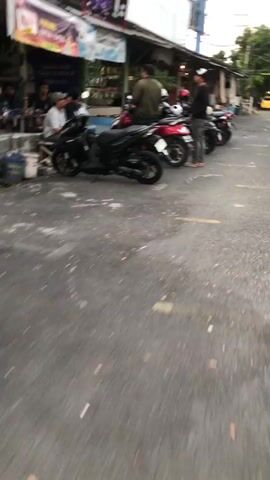 The video shows a street scene in front of what appears to be a small shop or restaurant. Several motorcycles are parked on the side of the road, with at least one red and one black scooter prominently visible. A few people are standing near the motorcycles and the shop entrance. One person, wearing a dark jacket and a baseball cap, is standing next to the red motorcycle. Another person in a light-colored jacket is standing further back, near the shop. The shop appears to have a sign that reads "WARUNG TANTE RIKA" and "KULINER". The ground is paved asphalt with some white markings. The lighting suggests it might be late afternoon or early evening. The camera is moving, causing some blur.