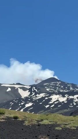 The video shows a volcanic mountain with patches of snow on its slopes. A plume of smoke is rising from the summit of the mountain, indicating volcanic activity. The sky is clear and blue, with a few white clouds. The camera appears to be stationary, capturing the plume of smoke as it drifts and expands. The lower portion of the video shows a grassy slope in the foreground. The overall scene is one of natural geological power.