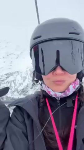 A woman wearing a dark ski helmet and goggles rides a ski lift on a snowy mountain. She is also wearing a black jacket and a pink floral neck covering. The camera is close to her face, and behind her, snow-covered mountains can be seen. The ski lift pole is visible on the left side of the frame. It appears to be a cloudy day.