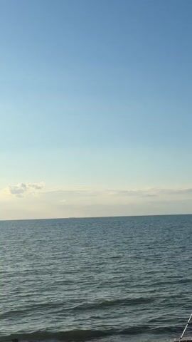 The video shows a calm body of water under a clear blue sky. Waves gently lap at a rocky shore in the foreground. A fishing rod is visible in the bottom right corner of the frame, suggesting someone is fishing. In the distance, a ship is barely visible on the horizon. The lighting indicates it is likely daytime, possibly late afternoon or early morning, given the soft quality of light. There are a few scattered clouds on the horizon.