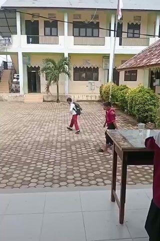 A video shows two young boys walking in a courtyard. One boy, wearing a red shirt and black pants, carries a piece of cardboard. The other boy, wearing a white shirt and red pants, has a backpack. They are walking away from a building that appears to be a school, with stairs leading up to a second story. The courtyard is paved with hexagonal tiles. A trash can is visible near a pillar in the foreground. The overall environment is outdoors, with some trees and other buildings visible in the background.