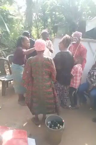 The video depicts a group of people gathered in what appears to be an outdoor area, possibly a yard or open space. There are several women and at least one man visible in the video. The women are dressed in various types of clothing, including dresses and patterned tops. One of them is wearing a head covering. The man is wearing a white shirt and jeans. The group appears to be engaged in some kind of activity, as some individuals are moving around and interacting with each other. The background environment includes foliage, suggesting a natural setting. Additionally, there is a container with bottles visible, and what seems to be a white board. The overall impression is of a social gathering or event taking place outdoors.