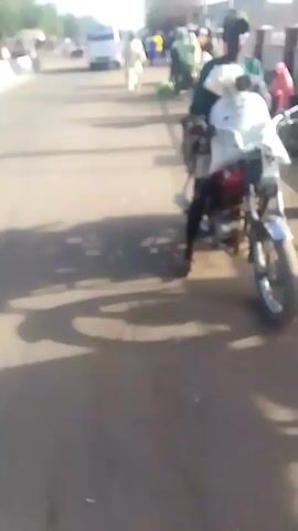 The video depicts a sunny outdoor scene on a paved surface. A man on a red motorcycle with a white bag is the focal point, passing by people selling watermelons spread out on the ground. The scene suggests a roadside market. Other individuals are visible in the background.