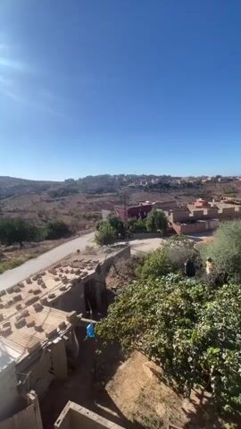 The video pans across a rural landscape under a clear blue sky. In the foreground, a partially constructed building with visible bricks is in the lower left corner. To the right, lush green foliage dominates, with a large banana tree in the center. Beyond this, a road winds through a village characterized by red-tiled roofs and sand-colored buildings. The village extends into the distance, meeting rolling, arid hills under the bright sunlight.
