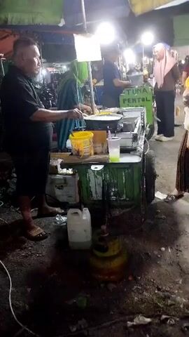 The video shows a nighttime scene at what appears to be a street food stall. There are several people present. In the foreground, a man wearing a black shirt and dark pants stands to the left, holding a spatula over a cooking surface. In front of him is a cart with various containers and cooking equipment. A yellow bucket is visible on the cart, along with a plastic container filled with liquid. To the right of the cart, a woman in a yellow shirt holds a plastic bag. Behind them, other individuals are visible, including a woman wearing a headscarf and another person in the background. The environment appears to be outdoors with multiple lights illuminating the area, suggesting a market or a busy street. There is a gas tank positioned on the ground in front of the cart. The ground is uneven and appears to be dirt or pavement with some debris. The cart itself is green and appears to be a mobile food vendor. A sign on the cart reads "TUNA PENTOL." The overall atmosphere is that of a lively street food setting at night.