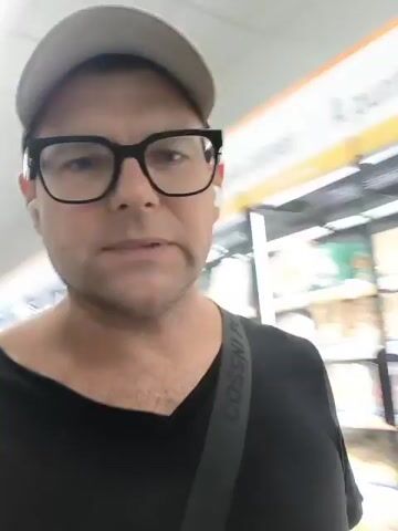 A medium close-up shot shows a man wearing a white baseball cap and black-rimmed glasses. He is wearing a black t-shirt and has a black strap across his chest. The man is looking directly at the camera and appears to be speaking. In the background, there are shelves stocked with various food items, including what appear to be packaged goods and bottles. The environment looks like a supermarket aisle with refrigerated display cases. A sign above the display case reads "A punt per a Salses" and "Zumos". The lighting is bright, typical of a retail setting. The video consists of a series of still frames, creating a stop-motion effect of the man's head moving slightly and his mouth opening and closing as if speaking.