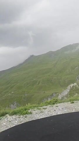 The video shows a view from a vehicle moving along a paved road. The road is covered in gravel in the foreground. In the background, there are green mountains with some rocky outcrops and patches of mist or fog. The sky is overcast. The camera appears to be mounted inside the vehicle, looking forward and slightly upwards.