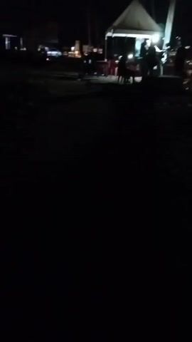 The video, shot at night, shows a poorly lit outdoor setting. A white tent is illuminated, with indistinct figures visible underneath.  Several chairs and tables are set up near the tent. The surroundings appear dark and undefined.
