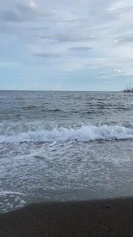 The video shows a view of a beach with dark sand and gentle waves rolling onto the shore. The sky is overcast with clouds. In the distance, a white boat is visible on the water. There appear to be two small figures swimming in the water between the shore and the boat. The video is a static shot from the perspective of someone standing on the beach looking out at the sea.
