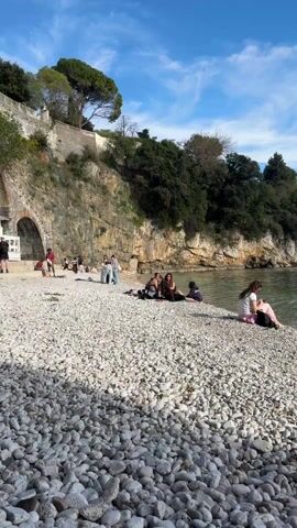 The video shows a pebble beach on a sunny day, with several people scattered along it. In the foreground, a young woman sits near the water's edge, looking out at the sea. Other people are gathered in small groups further back on the beach. The beach is composed of smooth, white pebbles. Behind the beach, a cliff rises, covered with trees and a stone wall. The water is clear and calm, with a few distant boats visible on the horizon. The sky is blue with scattered white clouds. The camera pans slightly to follow the coastline and then focuses on the sea. A person with dark curly hair walks into the scene, facing the water.