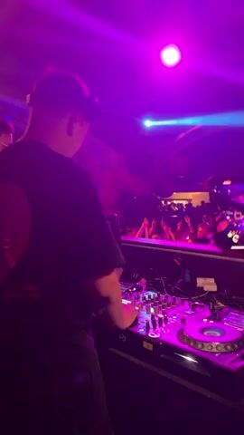 The video shows a DJ working at a party or club. The DJ is wearing a black shirt with a red graphic on the back and headphones over his ears. He is standing behind a DJ console, which is covered with various knobs, sliders, and turntables. The DJ is manipulating the equipment, presumably mixing music. In the background, a crowd of people can be seen dancing and enjoying themselves. The lighting in the scene is dynamic, with various colors flashing and changing, mostly blue and purple. There are also some large round decorations hanging from the ceiling.