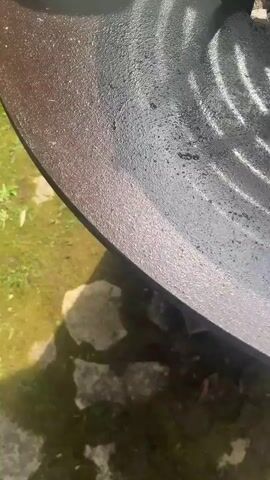 The video shows food being cooked on an outdoor grill. A large piece of meat sits on a raised, circular grill grate. Below the grate, on a flat surface, are slices of toasted bread and strips of cooked meat and sausages. The grill is located outside on a grassy area with some stone pavers. The scene is well-lit.
