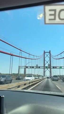 The video shows a first-person perspective from inside a vehicle driving across a suspension bridge. The bridge has red cables and towers, and there are many cars on the bridge in front and to the sides. Traffic signals are visible, indicating traffic is moving. The sky is clear and blue. In the distance, a tall statue or monument is visible on the left side of the bridge. The interior of the car is partially visible, including the dashboard and windshield.