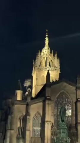 The video shows St. Giles' Cathedral in Edinburgh, Scotland, at night. The building is illuminated with bright yellow light. A statue stands in front of a large window in the lower right part of the frame. White smoke drifts upwards from behind one of the cathedral's towers. A bagpipe can be heard playing in the background.