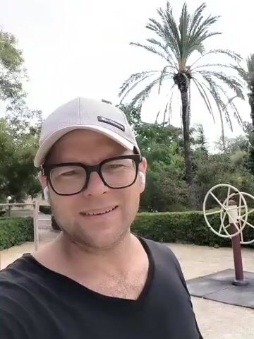 A man wearing glasses and a baseball cap is standing outside, smiling at the camera. He is wearing a black t-shirt. In the background, there are palm trees, bushes, and some outdoor exercise equipment. Another person can be seen in the distance using one of the machines. The man is speaking in Russian, describing the beauty of the place and that he is exercising.
