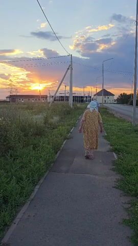 An elderly woman walks down a sidewalk at sunset. She is wearing a yellow patterned dress and a light blue headscarf. Behind her, a man walks in the same direction. The sky is filled with orange and purple clouds as the sun sets behind a row of buildings. The sidewalk is bordered by green grass. Power lines run across the sky.