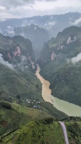 The video shows an aerial view of a wide, muddy river flowing through a lush green valley flanked by steep, tree-covered mountains. The sky is overcast with clouds obscuring the mountain peaks in the distance. Several boats are visible on the river, leaving white wakes as they move downstream. In the foreground, on the left side of the riverbank, a small cluster of houses forms a village nestled into the hillside. A winding road with a striped barrier is visible on the right side of the frame, descending towards the valley floor. The camera pans slightly, offering a sweeping panorama of the dramatic landscape.